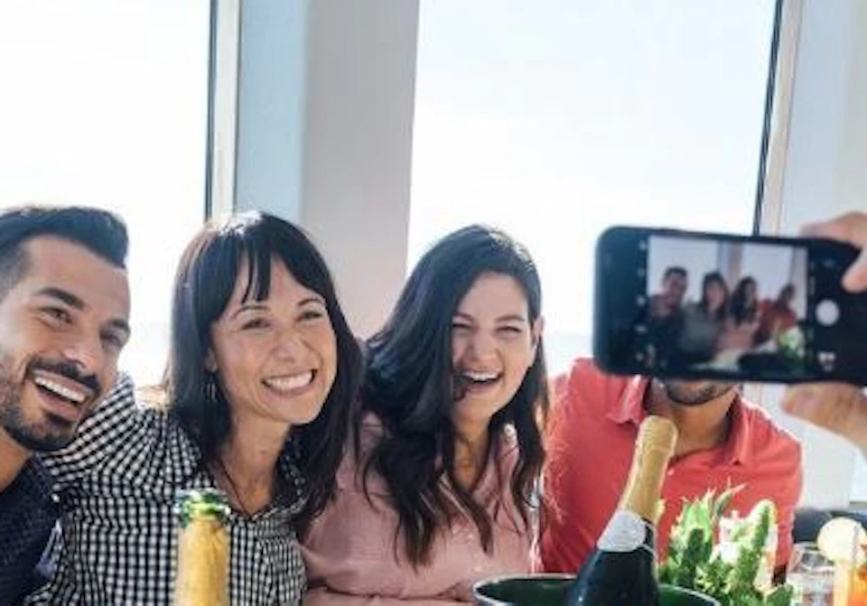 A group of friends smiling at a table with food and drinks, while someone takes a photo with a smartphone.