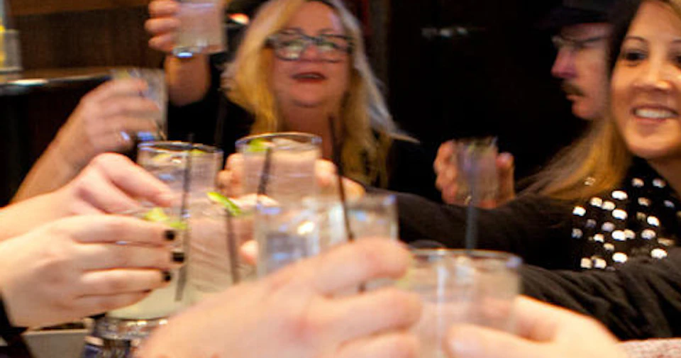 A group of people at a wooden table toasting with drinks, with food and condiments placed in front of them.