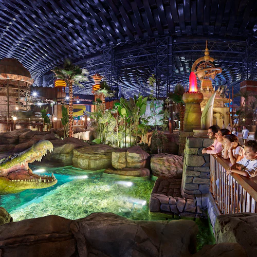 A family observes an animatronic crocodile in a brightly lit indoor amusement park with tropical-themed decor.