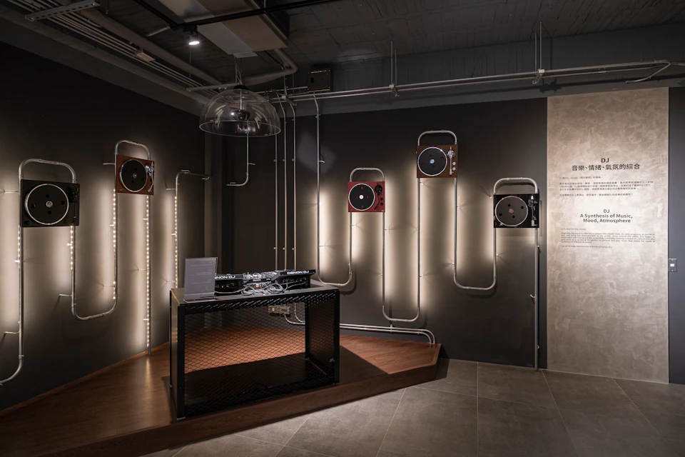 A modern interior space with a DJ setup on a metal table and wall-mounted speakers integrated into a pipe design.