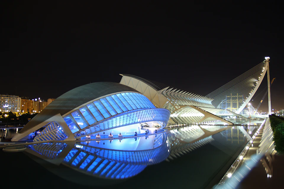 Modern, illuminated architecture with curved, futuristic designs is reflected in a calm body of water against a dark night sky.