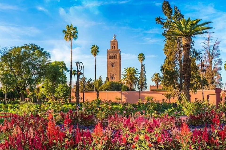 A tall, ornate tower rises above vibrant gardens with pink flowers and tall palm trees under a blue sky.
