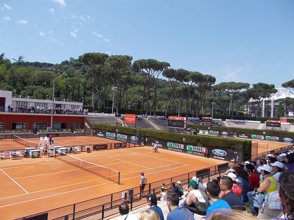 一场网球比赛正在室外粘土球场上进行，看台上有观众，后面是树木。