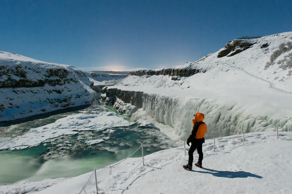 A person in winter clothing stands by a rope barrier, overlooking a frozen waterfall and snowy landscape under a clear sky.