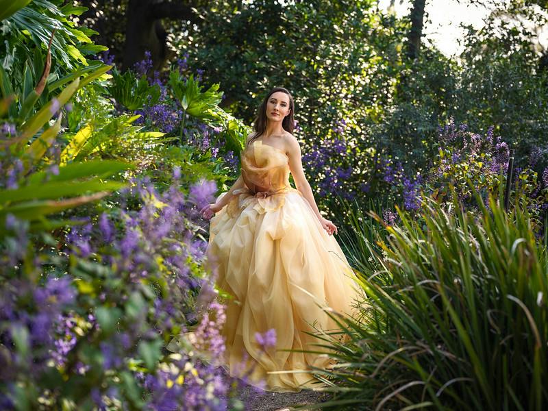 A woman in a flowing yellow gown stands in a lush garden surrounded by green foliage and purple flowers.