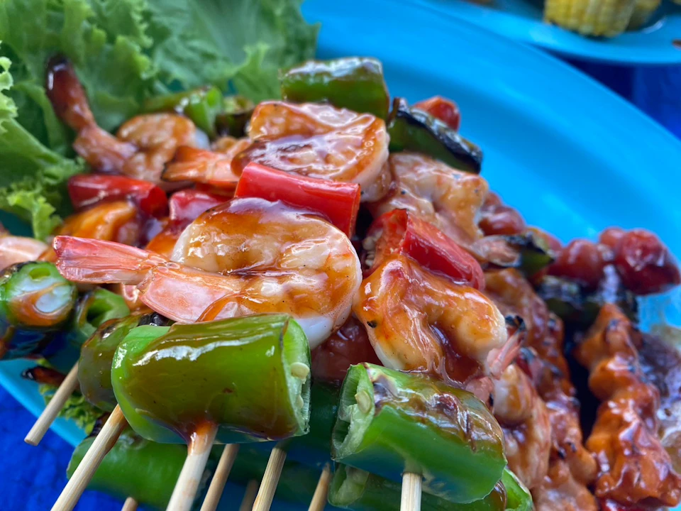 Close-up of grilled shrimp and vegetables on skewers, glazed with sauce, served on a blue plate with a lettuce garnish.