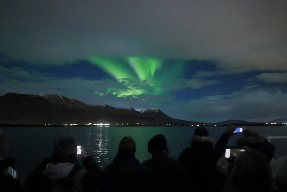People observing and photographing the green Northern Lights over a calm lake, with mountains in the background.