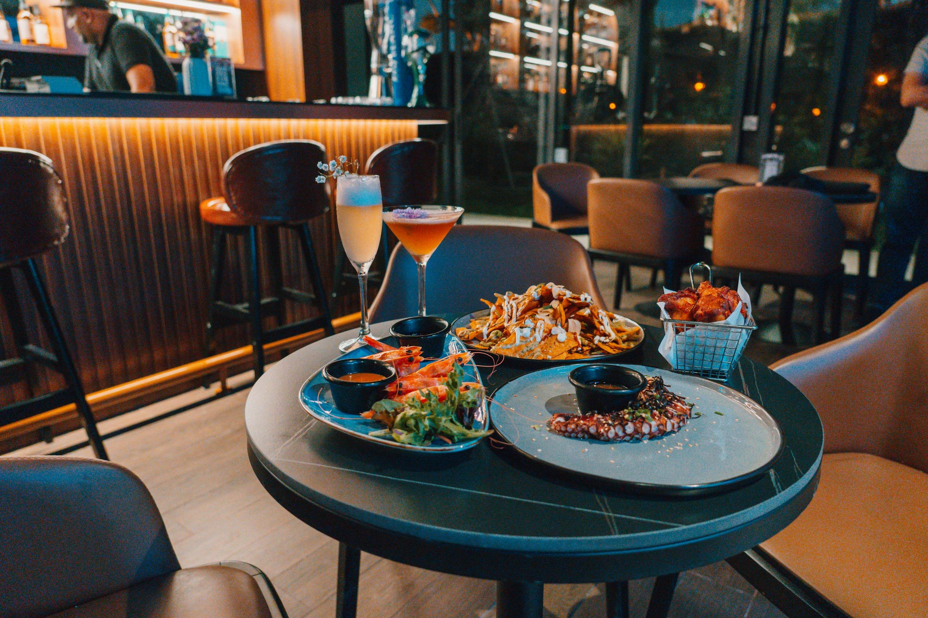 A table in a modern restaurant with dishes including seafood, fries, and cocktails, surrounded by seating and a bar area.