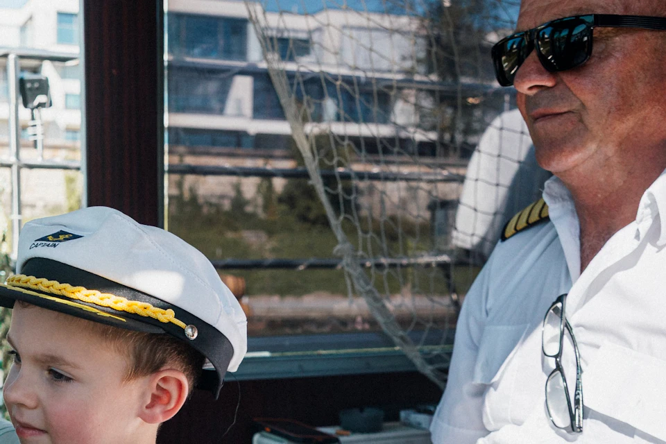 A young boy wearing a captain's hat steers a boat while an older man in a captain's uniform sits beside him.