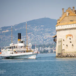 A steamboat cruises near a stone castle with a mountainous landscape in the background.