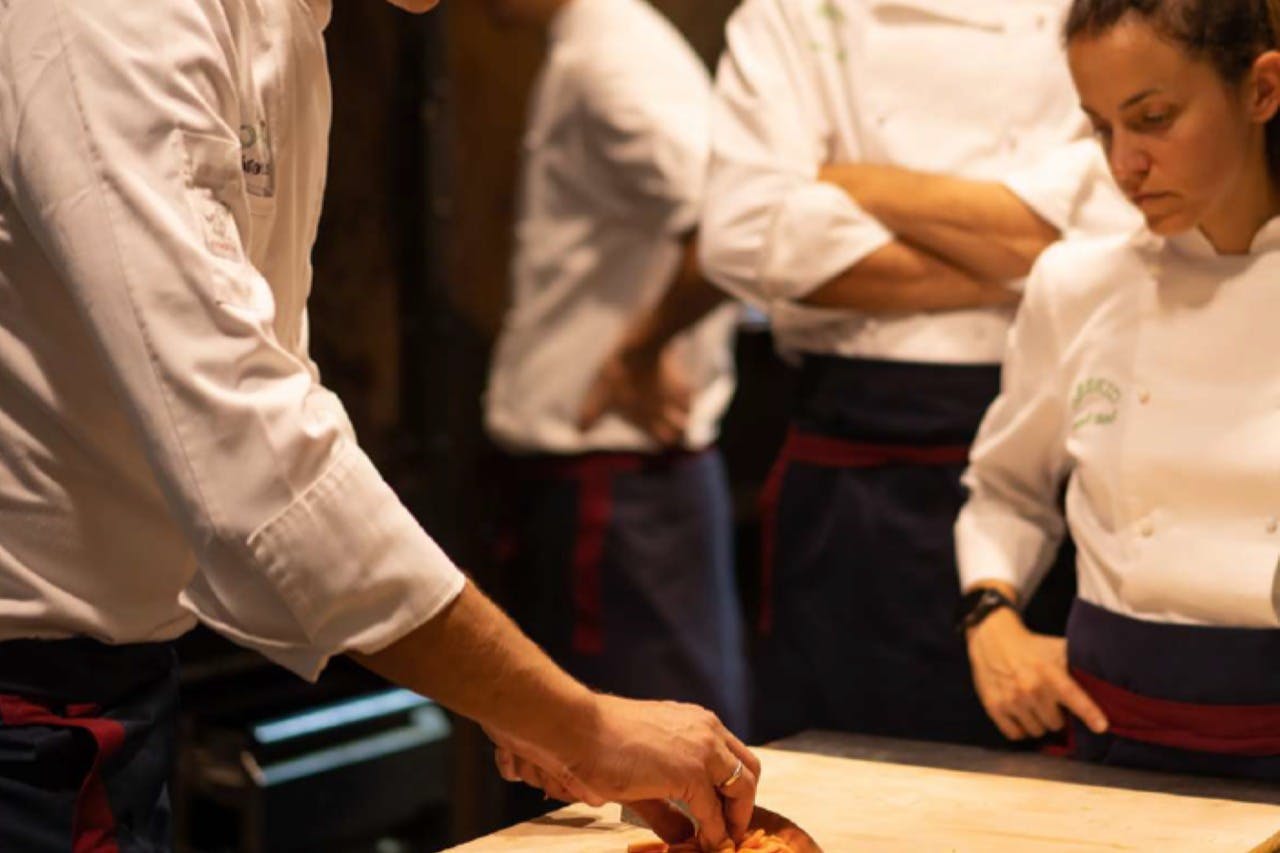 Chefs in white uniforms and blue aprons stand around a table, intently watching one chef slice a piece of food.