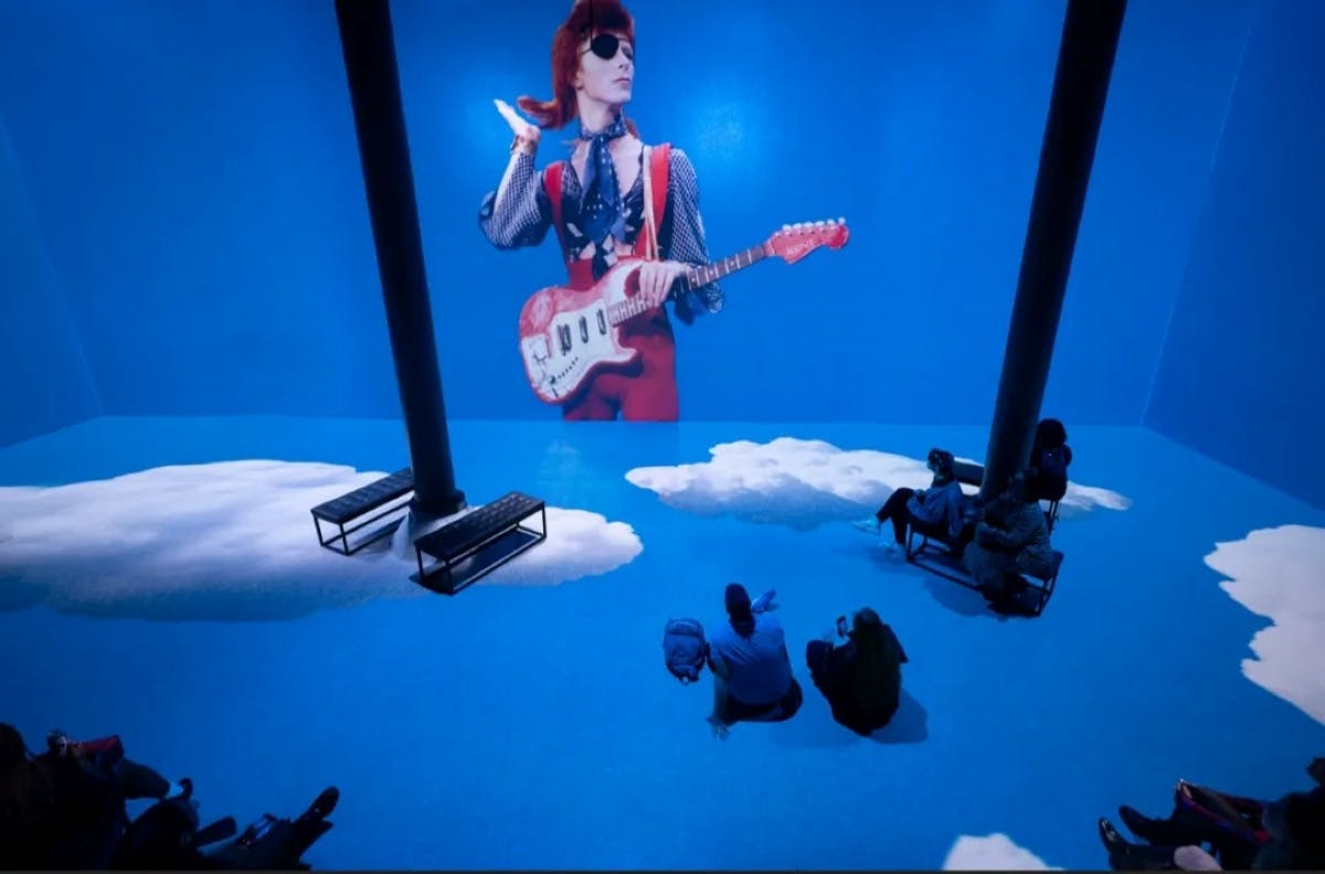 人们坐在一个大屏幕的蓝色房间里，展示了一位演奏电吉他的音乐家。 有长凳和柱子。