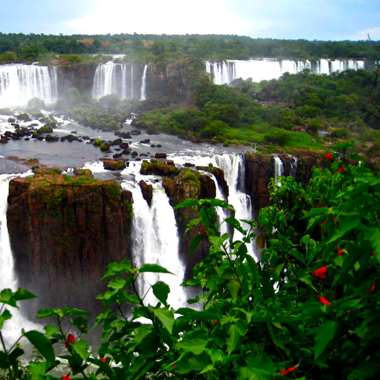 Multiple waterfalls cascade over cliffs surrounded by lush green vegetation and vibrant red flowers.