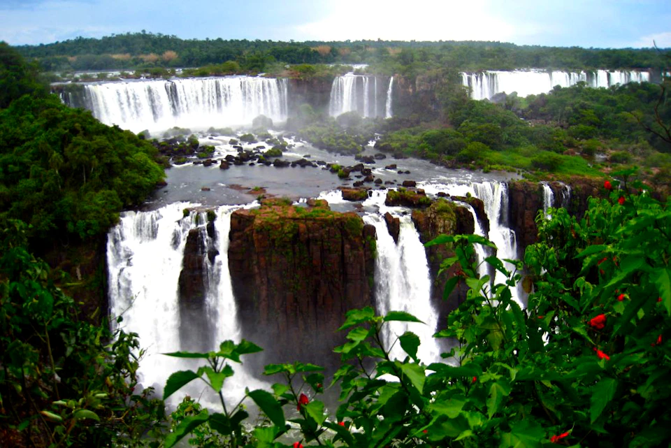 Multiple waterfalls cascade over cliffs surrounded by lush green vegetation and vibrant red flowers.