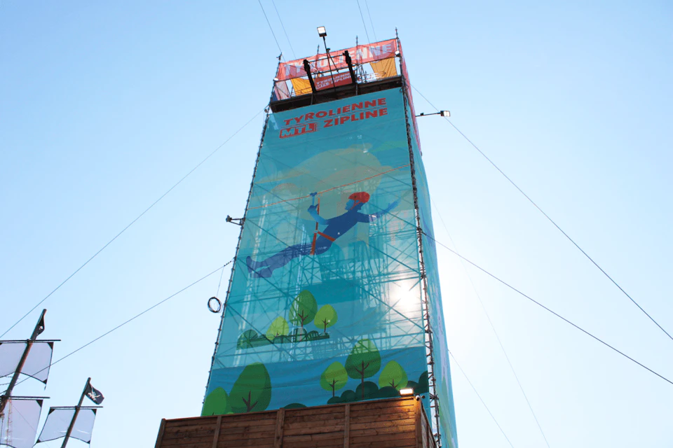 A tall structure with a zipline setup. The sign reads “Tyrolienne / Zipline” and it features an illustration of a person ziplining.