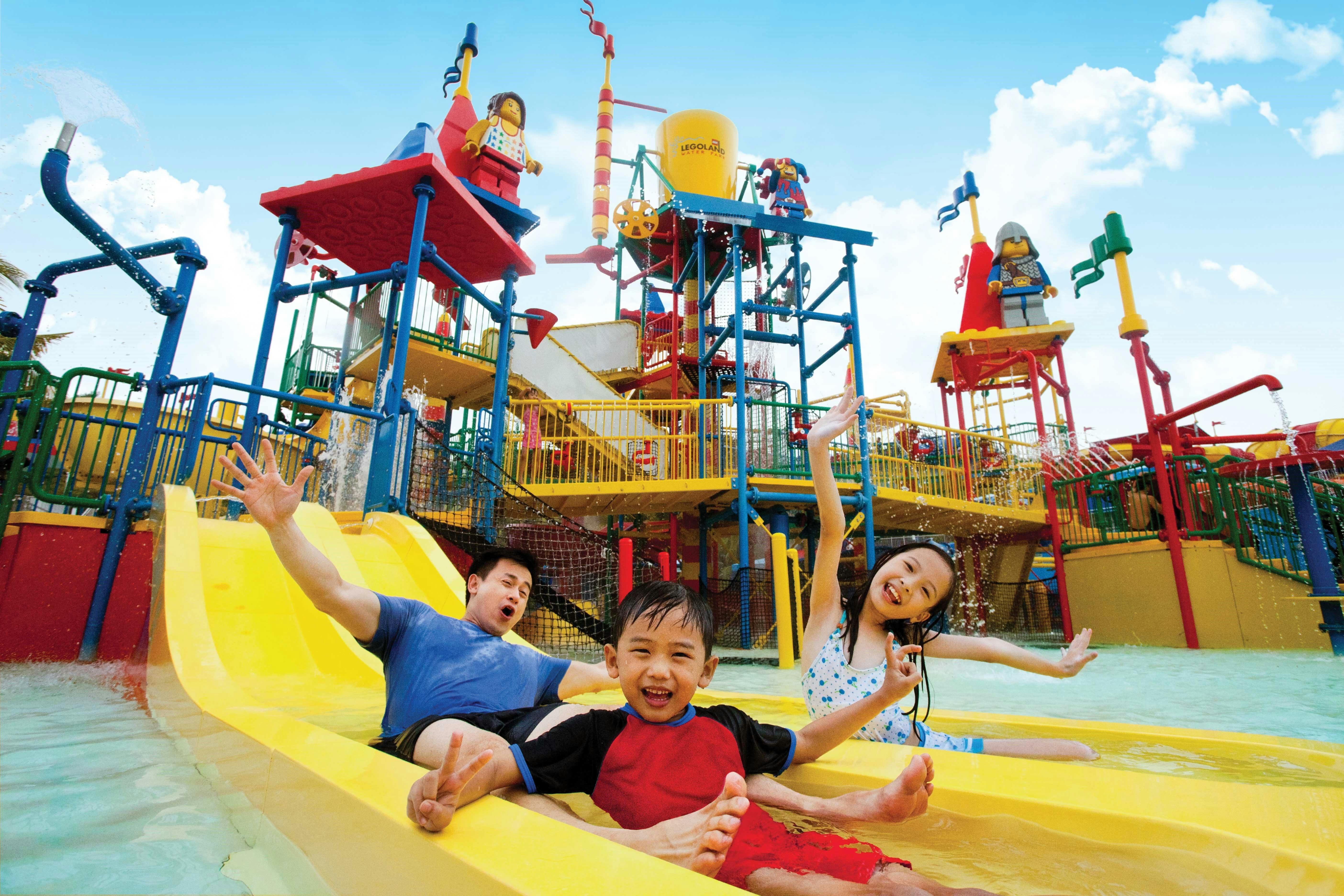A family of three joyfully slides down a yellow water slide at a colorful waterpark under a bright blue sky.