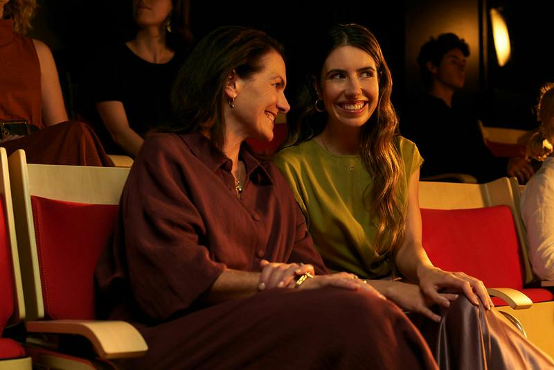 Two women smiling and sitting together in a warmly lit room, with others blurred in the background.