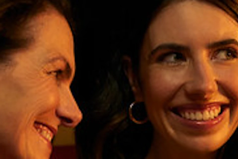 Two women smiling and sitting together in a warmly lit room, with others blurred in the background.