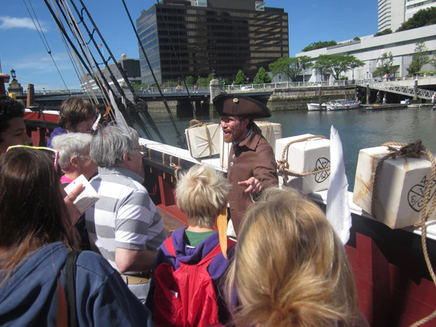 A man dressed as a pirate speaks to a group of people on a historic ship docked by a waterfront with buildings in the background.