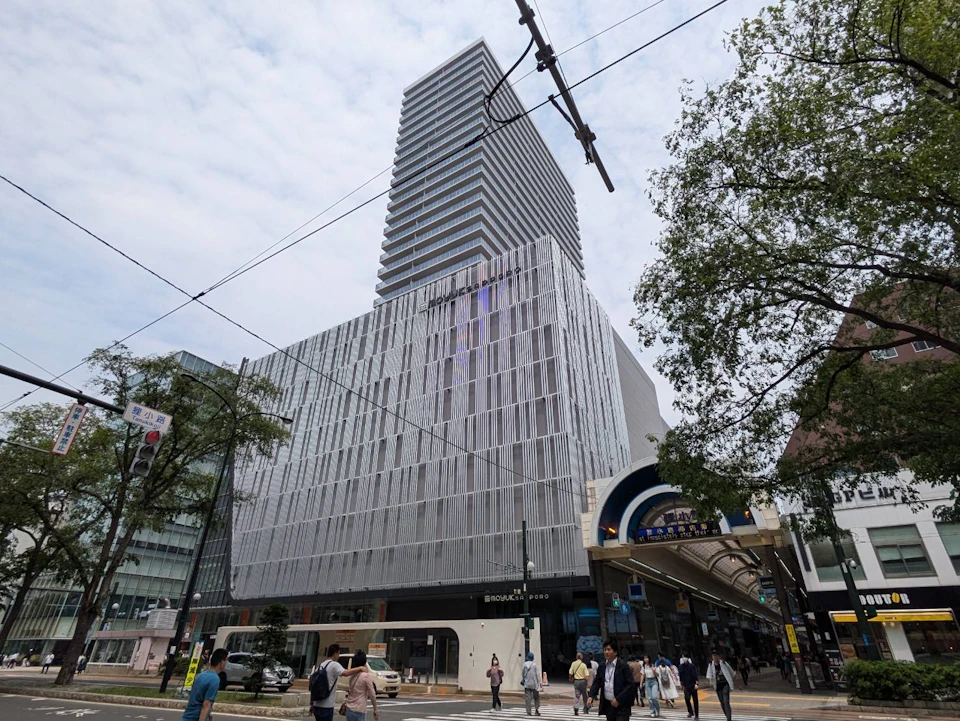 A modern multi-story building with a grid-like facade and a street scene with pedestrians and trees in the foreground.