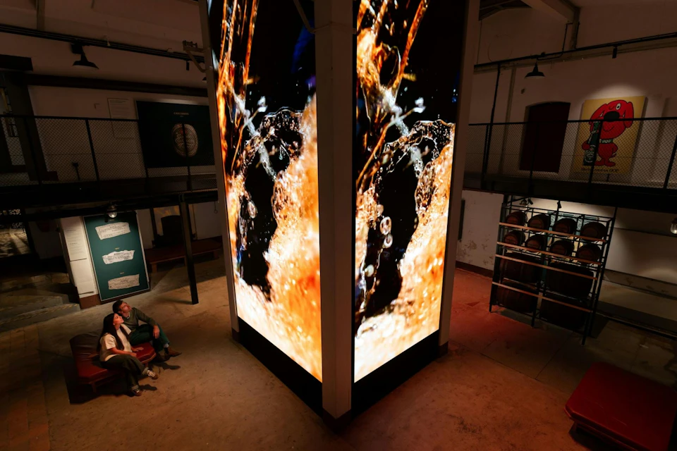 A large vertical digital screen displays an artistic image of splashing liquid. Two people sit on a bench nearby in a dimly lit room.