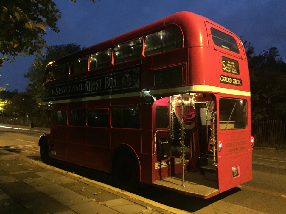 A red double-decker bus with "Ghost Bus" signage, parked at dusk with its door open and interior lights on.