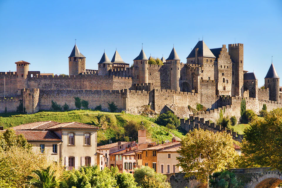 A historical castle with multiple towers overlooks a village with colorful houses and green foliage under a clear blue sky.