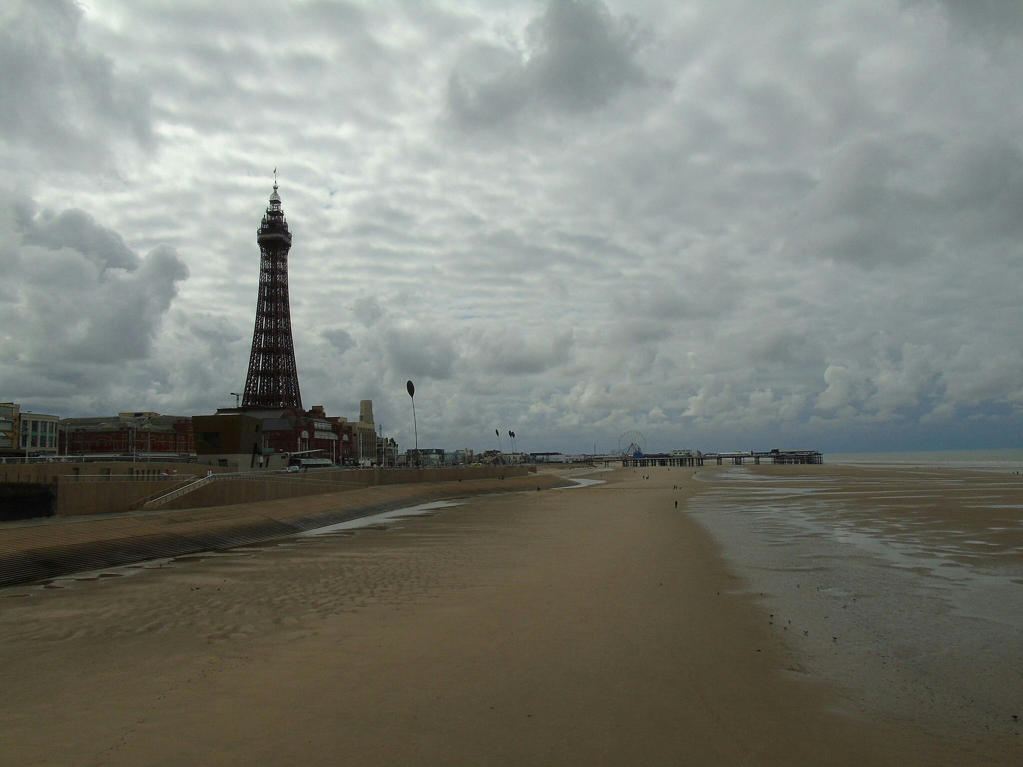 Una spiaggia con sabbia bagnata, una ruota panoramica lontana e un'alta torre sotto un cielo nuvoloso. Poche persone sono visibili nella scena.