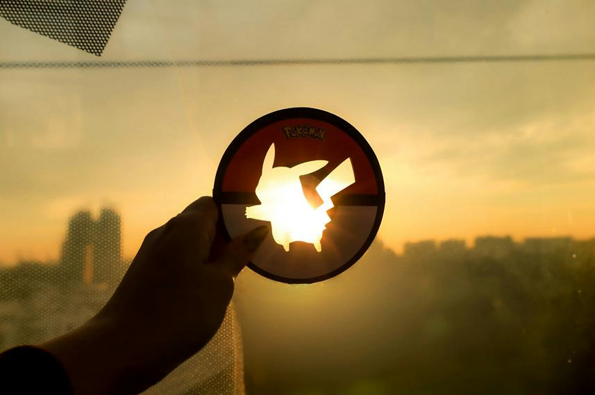 A hand holds a circular Pokémon-themed object with a Pikachu cutout against a sunset-lit background.
