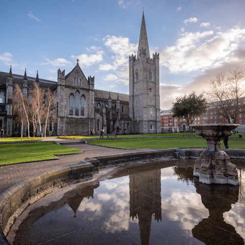 En historisk stenkatedral med en hög spiral bredvid en reflekterande fontän, omgiven av gröna gräsmattor och några personer under en blå himmel.