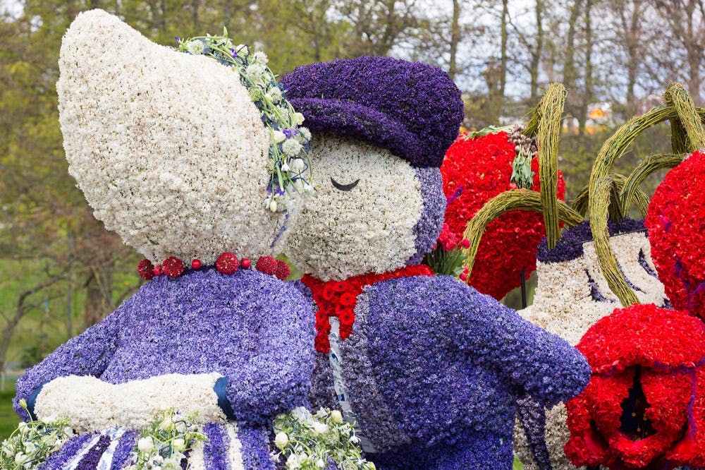 Δύο ανθρώπινες φιγούρες φτιαγμένες εξ ολοκλήρου από λουλούδια, ντυμένες σε μοβ και λευκό, παρουσιάζονται σε μια στάση φιλιού, σε εξωτερικούς χώρους.