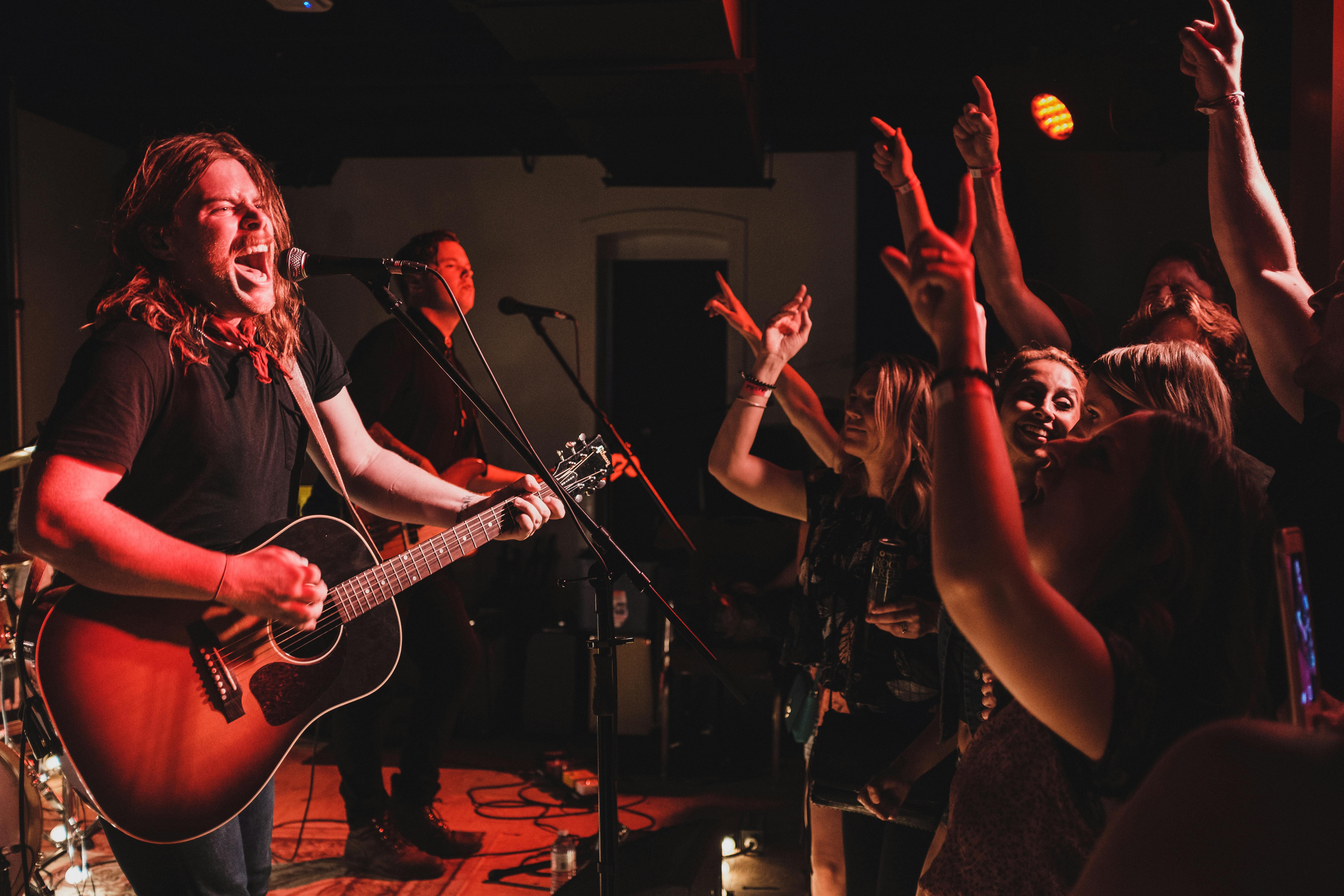 Un musicista che canta con passione e suona la chitarra sul palco con una band, mentre il pubblico entusiasta applaude.