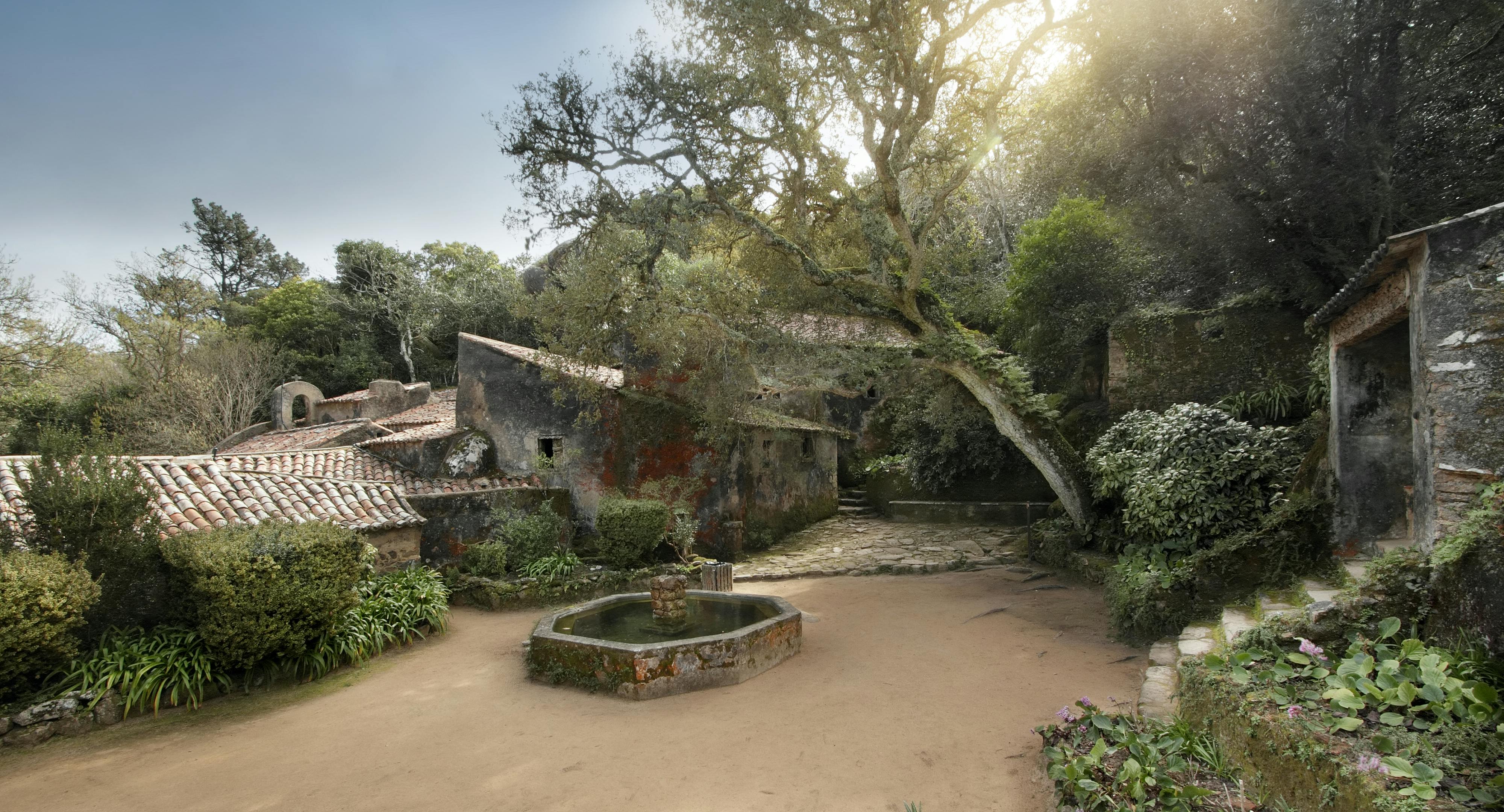 Cortile in pietra con un'antica fontana, circondato da alberi ed edifici rustici con pareti ricoperte di muschio e tetti di tegole.