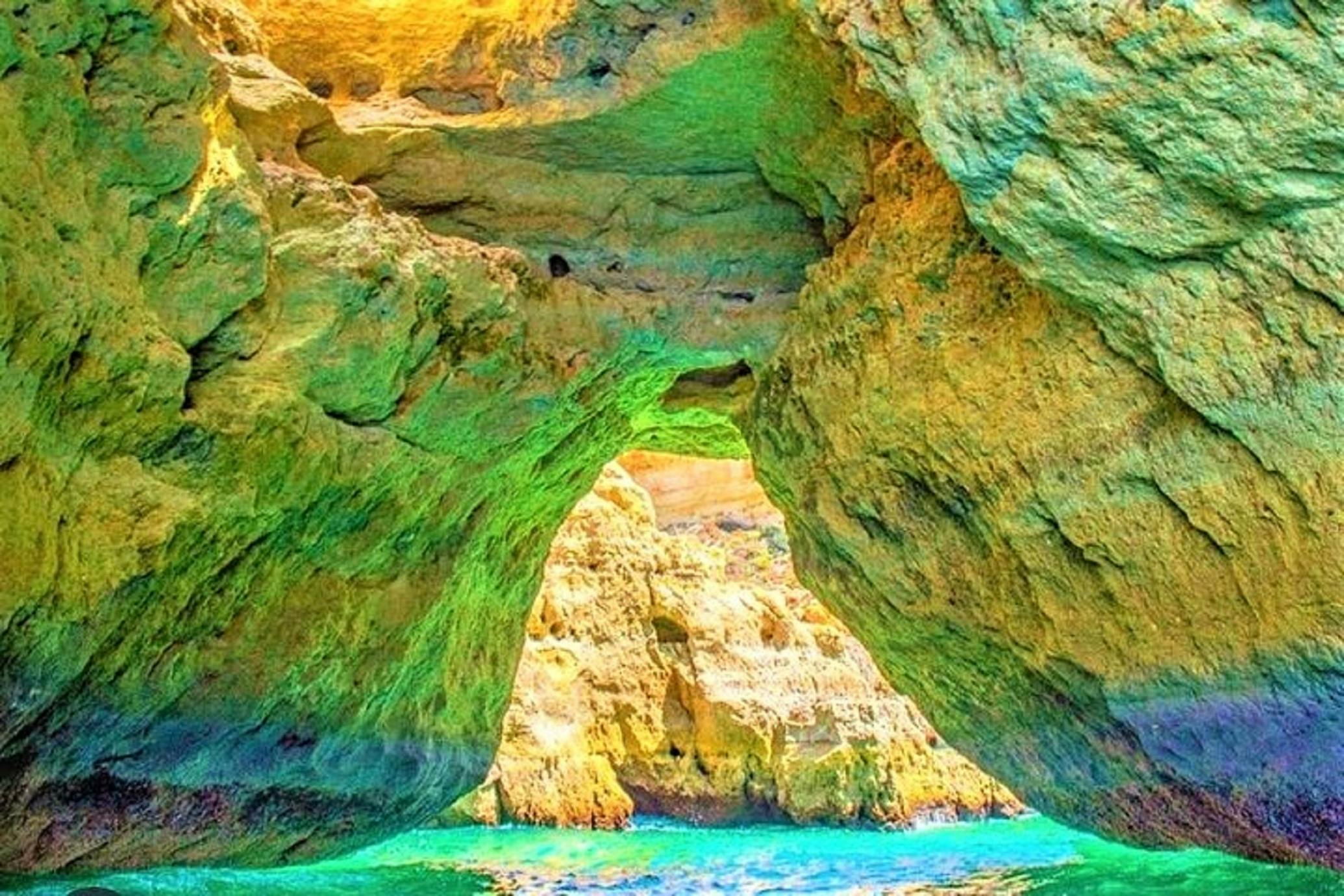 Sunlit cave with vibrant, multicolored rock formations and blue water at the base. Natural rock arch in the center.