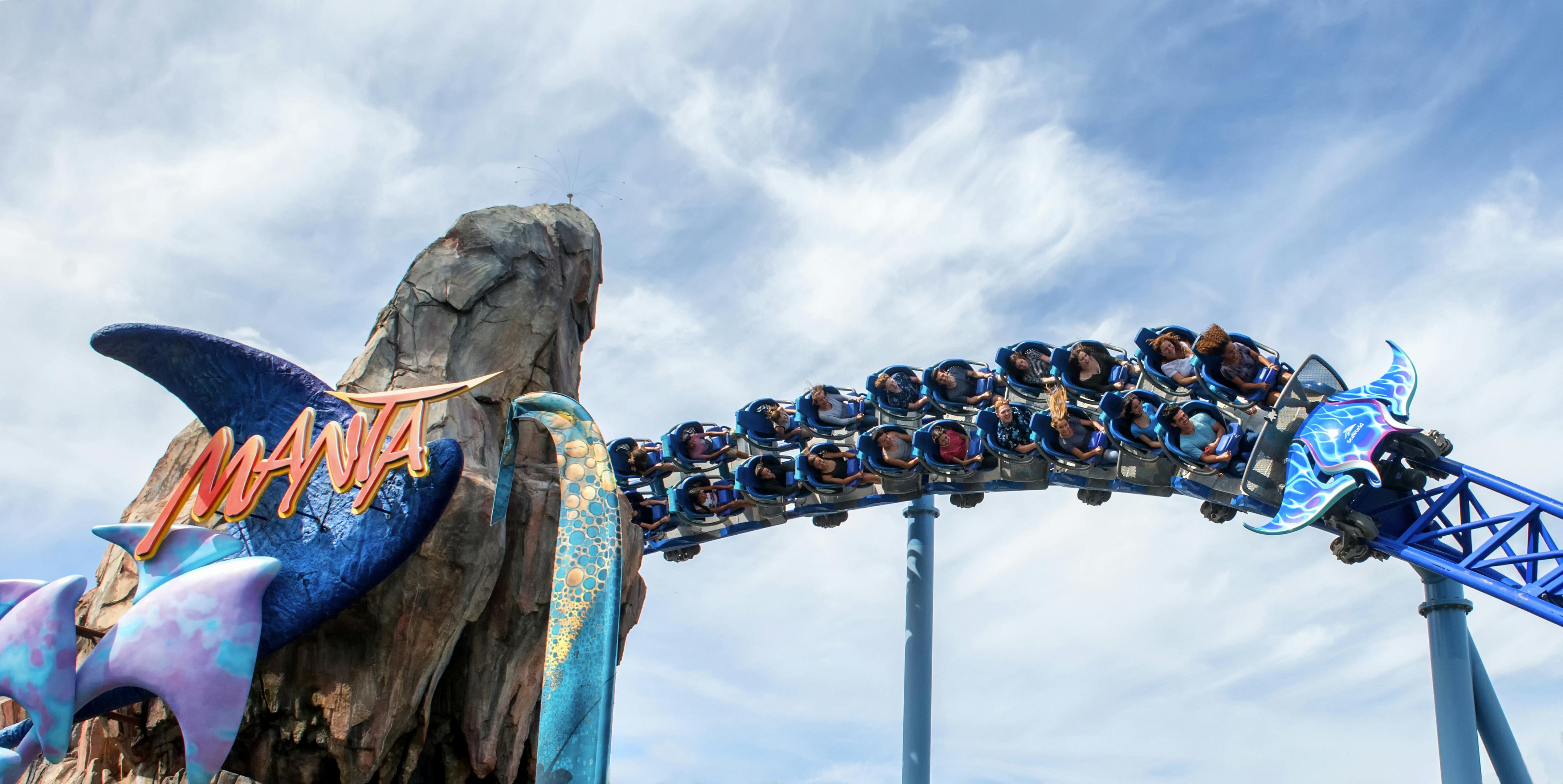 Άνθρωποι καβαλικεύουν ένα μπλε τρενάκι του λούνα παρκ, γυρνώντας γύρω από μια βραχώδη δομή στολισμένη με θεματικές διακοσμήσεις, κάτω από έναν εν μέρει συννεφιασμένο ουρανό.