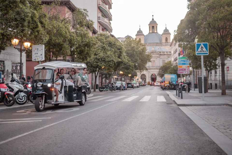 A busy street lined with trees and buildings, featuring a tuk-tuk with passengers and a historic domed building in the background.