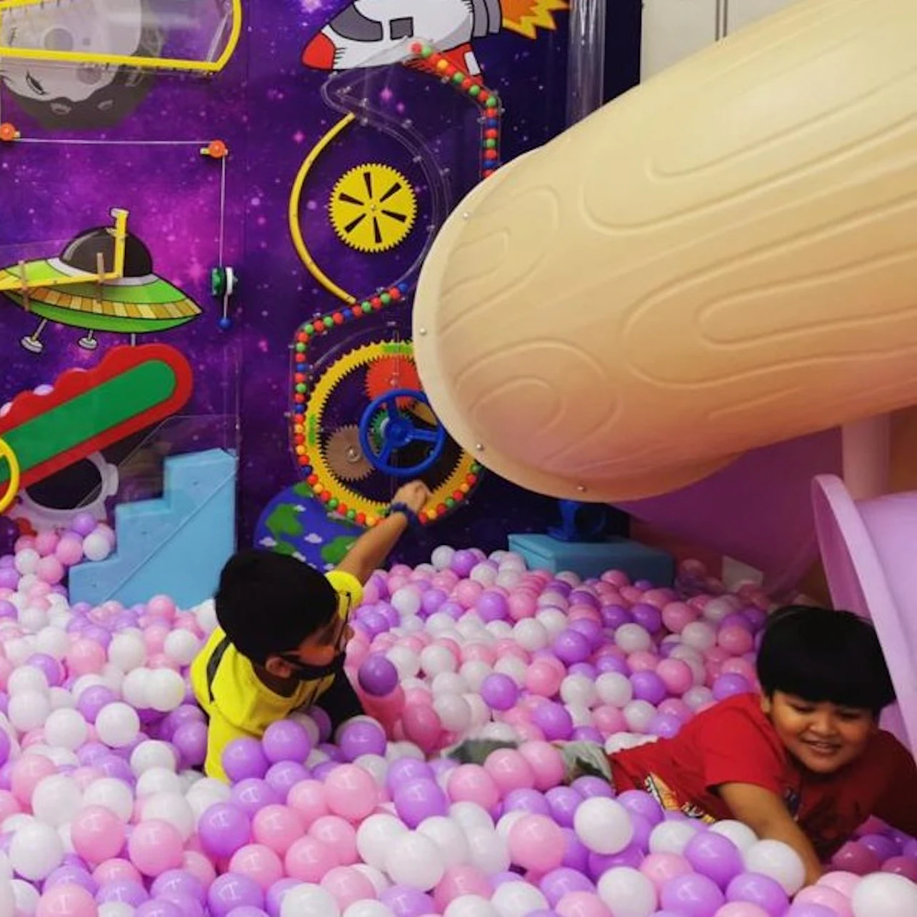 Two children playing in a colorful ball pit with a space-themed background and a slide.