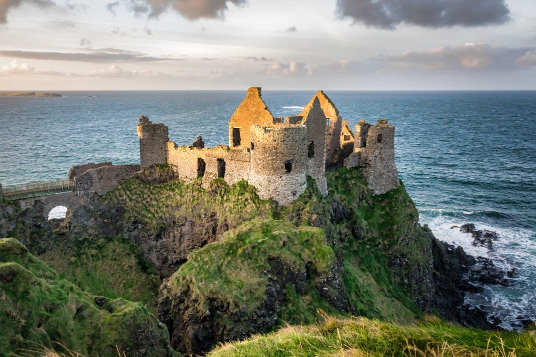 Les ruines d'un château historique en pierre sont perchées au sommet d'une falaise herbeuse surplombant l'océan sous un ciel nuageux.