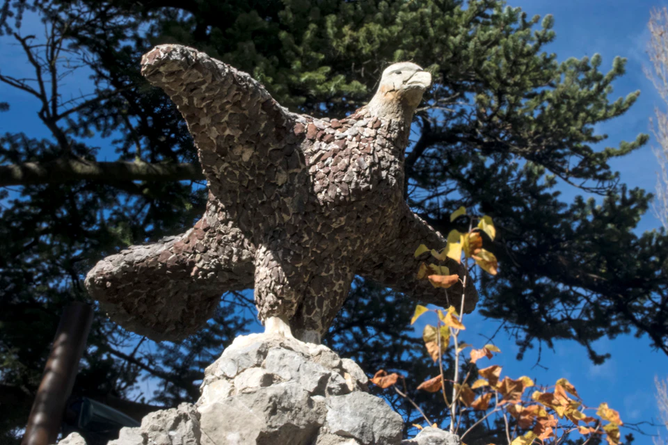 Stone sculpture of an eagle perched on a rock, with a background of trees and a clear blue sky.