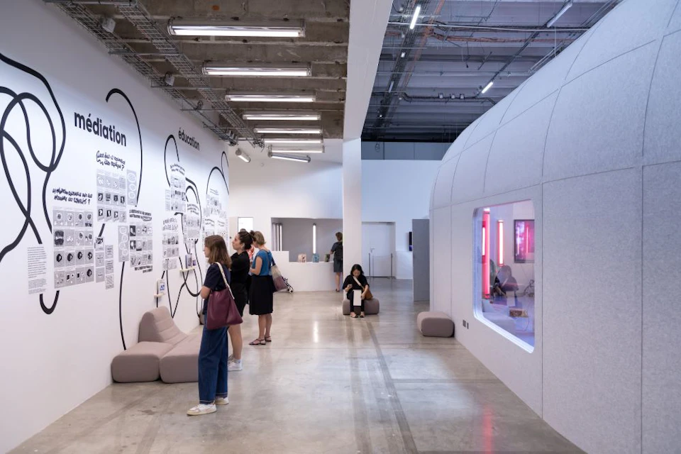 A modern exhibition space with people viewing wall displays and sitting on minimal furniture, featuring bright lighting.