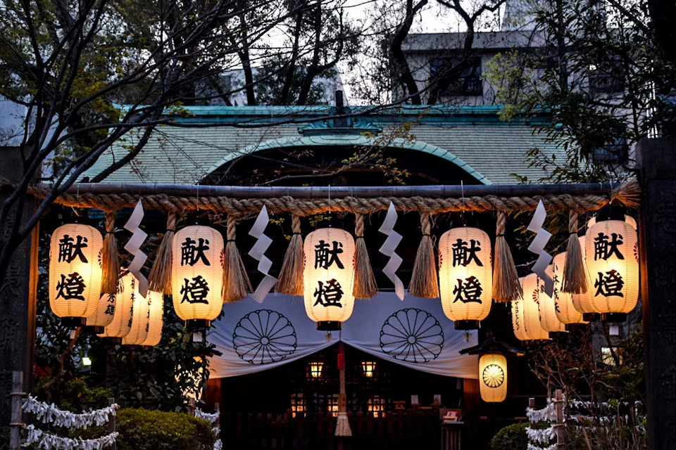 Hanging lanterns with Japanese characters and white paper decorations in front of a traditional building surrounded by trees.