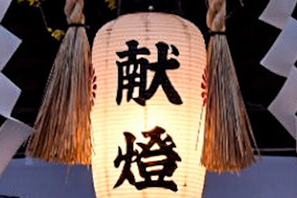 Hanging lanterns with Japanese characters and white paper decorations in front of a traditional building surrounded by trees.