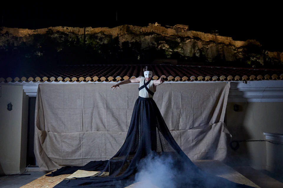 A performer in a white mask and black dress stands on a stage with smoke, with an ancient lit structure atop a hill in the background.