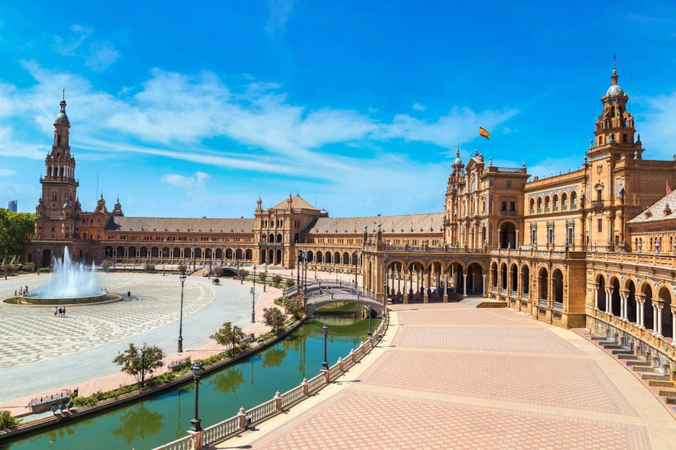 Panoramic view of Plaza de España in Seville, Spain, featuring curved buildings, a central canal, and a large open plaza under a blue sky.
