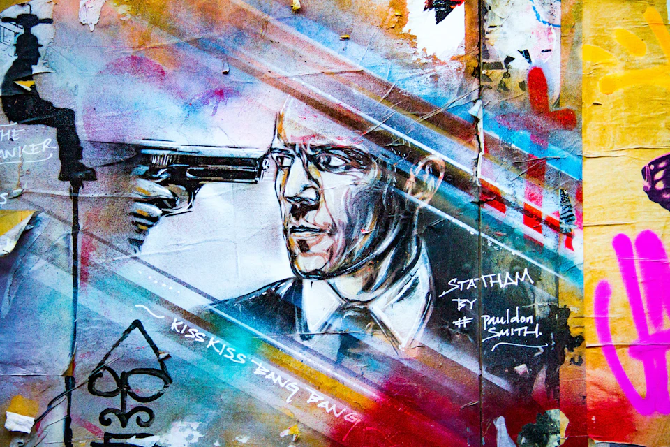 A mural depicts a man in a suit pointing a gun at his head. Text nearby reads: "Statham," "BY #pauldonsmith," and "Kiss Kiss Bang Bang.”