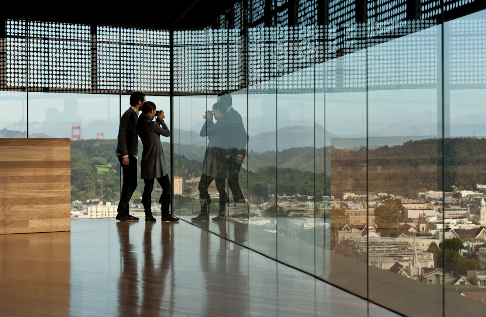 Two people in business attire look out through floor-to-ceiling windows with a city and landscape reflected in the glass.