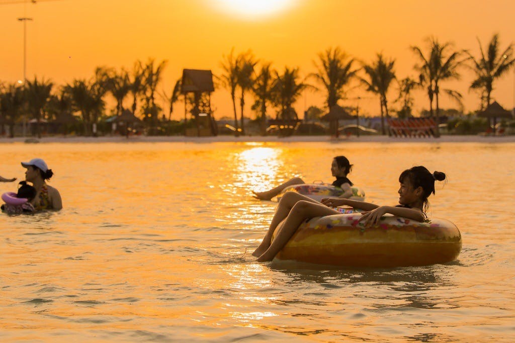 Two people relaxing on inflatable rings in water at sunset, with palm trees lining the background.