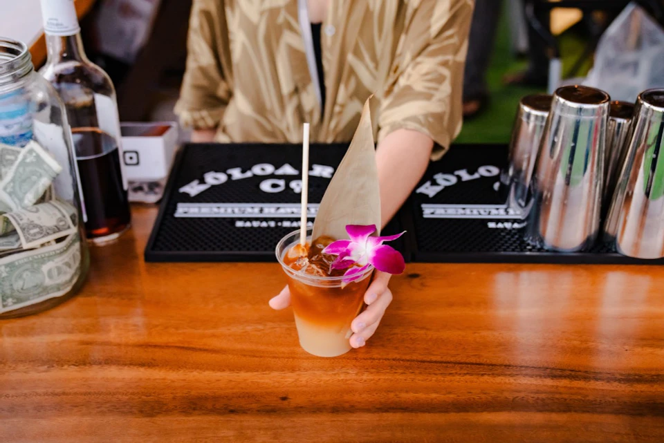 A person wearing a tan shirt holds a tropical cocktail garnished with an orchid and a leaf at a wooden bar counter.