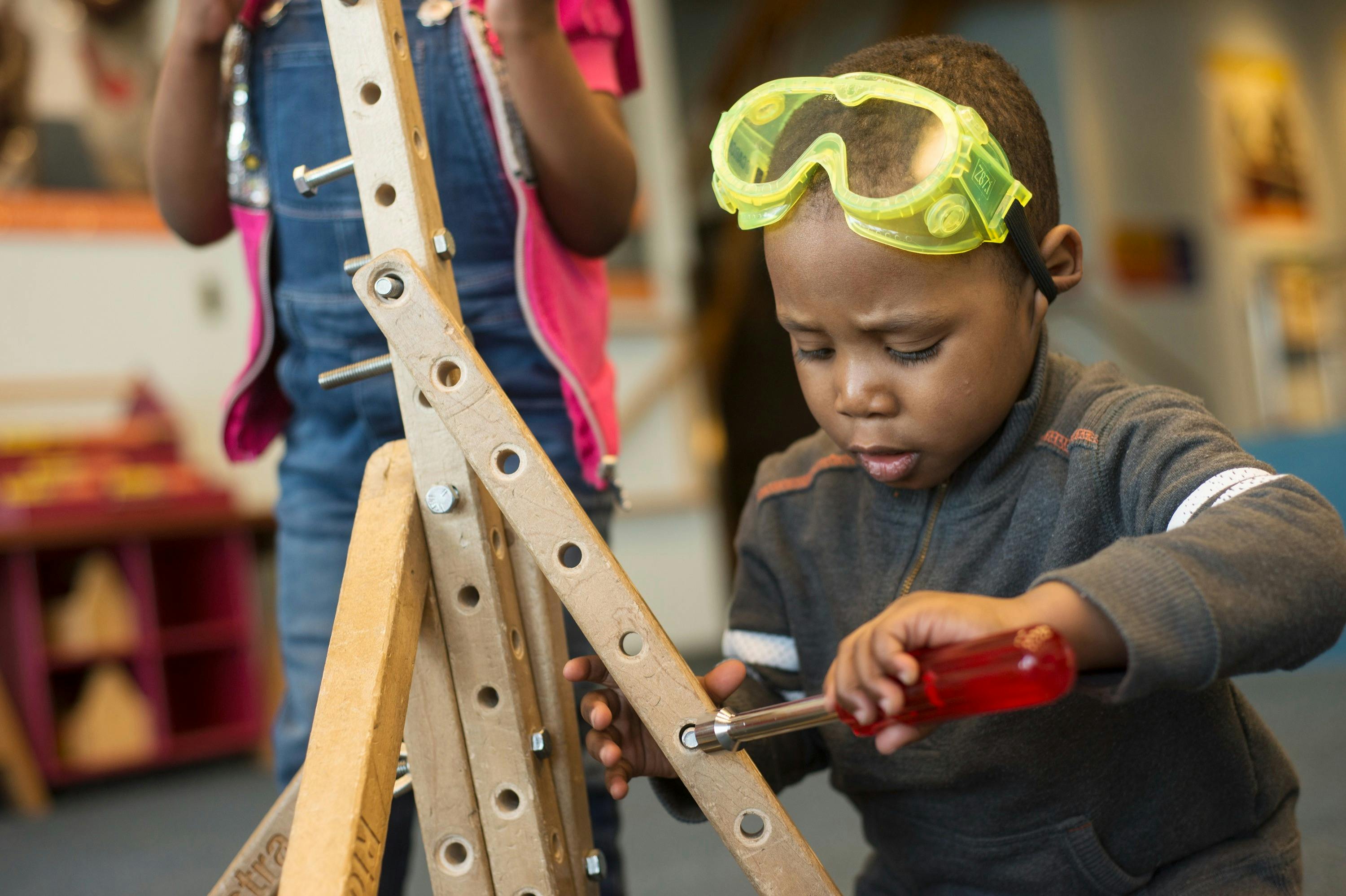 A child wearing safety goggles uses a screwdriver to work on a wooden structure in an indoor environment.