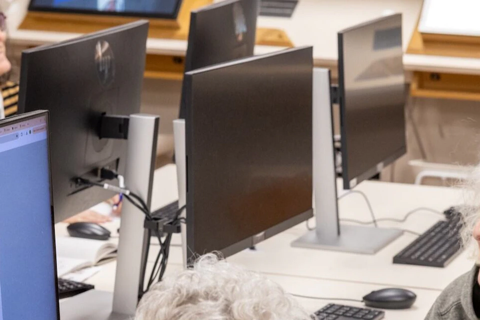 A group of people sitting at computer workstations in a library, engaged in an activity on the screens.